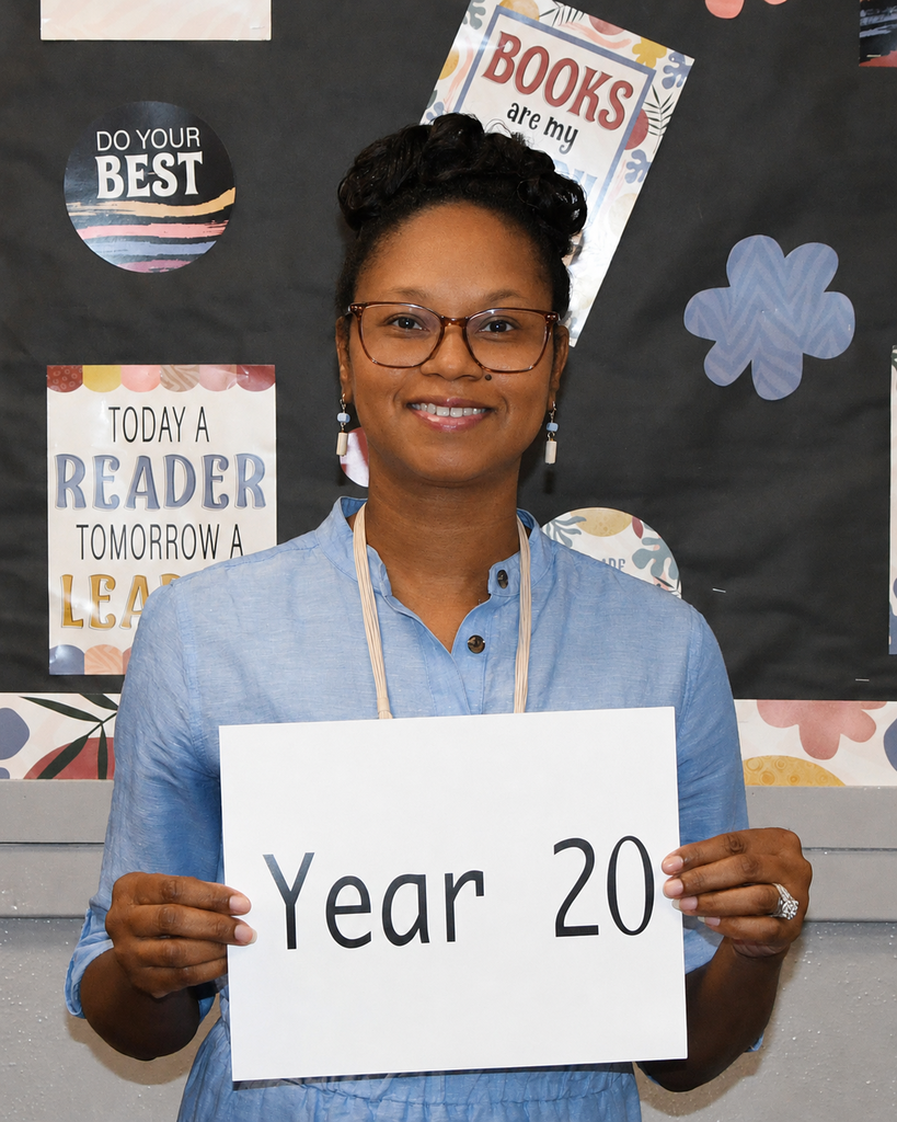 Woman in blue shirt holding a white sign that reads "Year 20" in a classroom with posters on the wall.