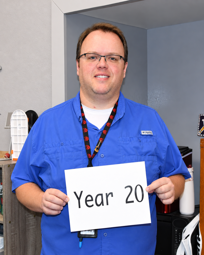 Man in blue shirt and glasses holding a white sign that says "Year 20". Behind him is a room with gray walls.