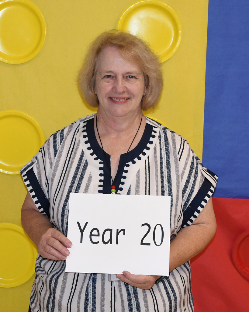 A woman in a striped shirt holds a white sign that says "Year 20" in front of a colorful background.