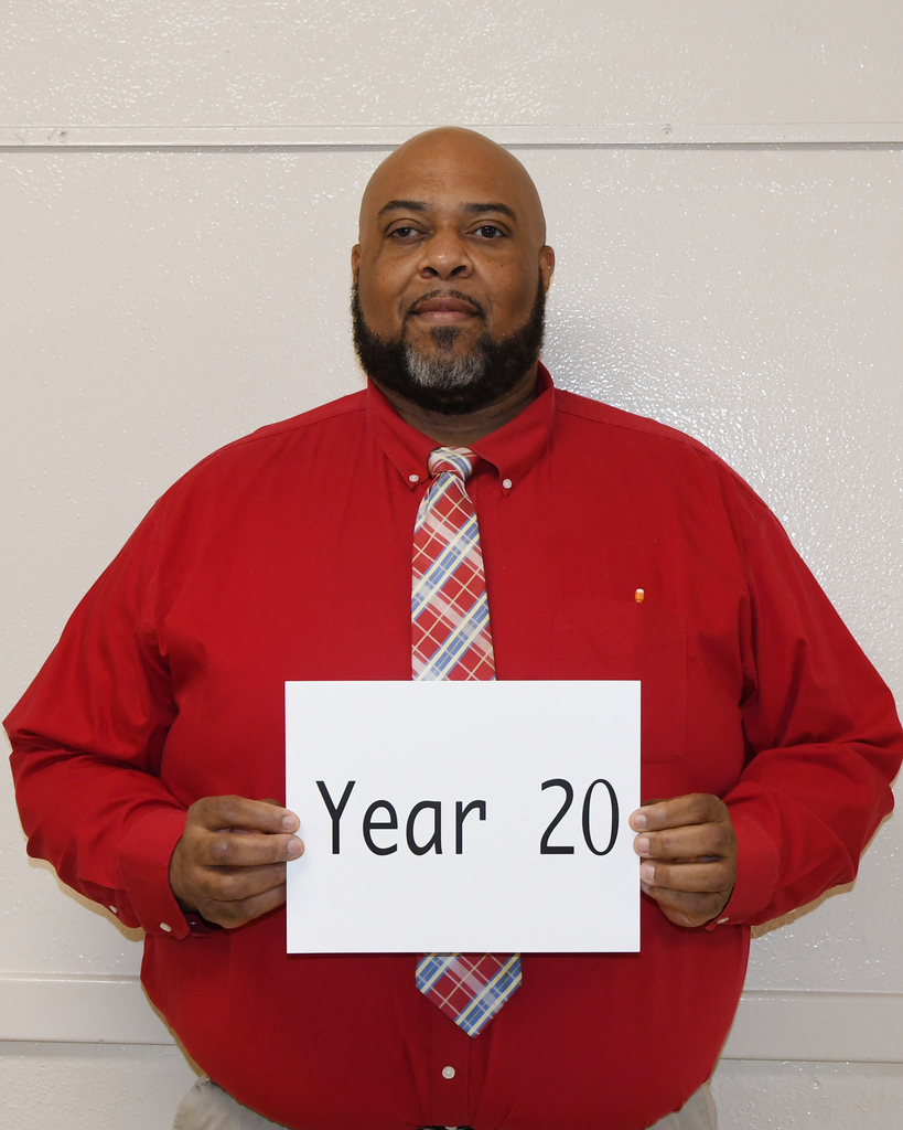 A man in a red shirt and tie holds a white sign reading "Year 20" in front of a white wall.
