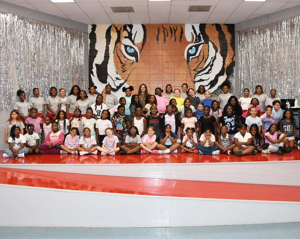 Group photo of children and adults on a stage in front of a large tiger mural.