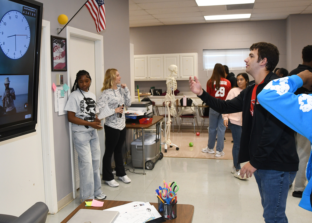 A classroom with a skeleton model, large screen, and several people standing or walking. Some are engaged in conversation.