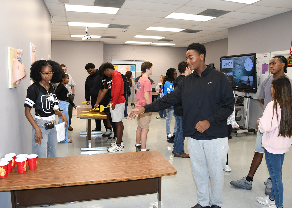 People interact in a room with tables, red cups, and monitors. Some wear jackets and sneakers.