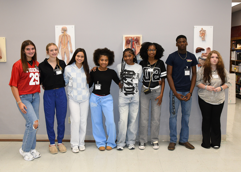 Seven young adults stand in a line against a wall with medical posters. They wear varied colored tops and pants.