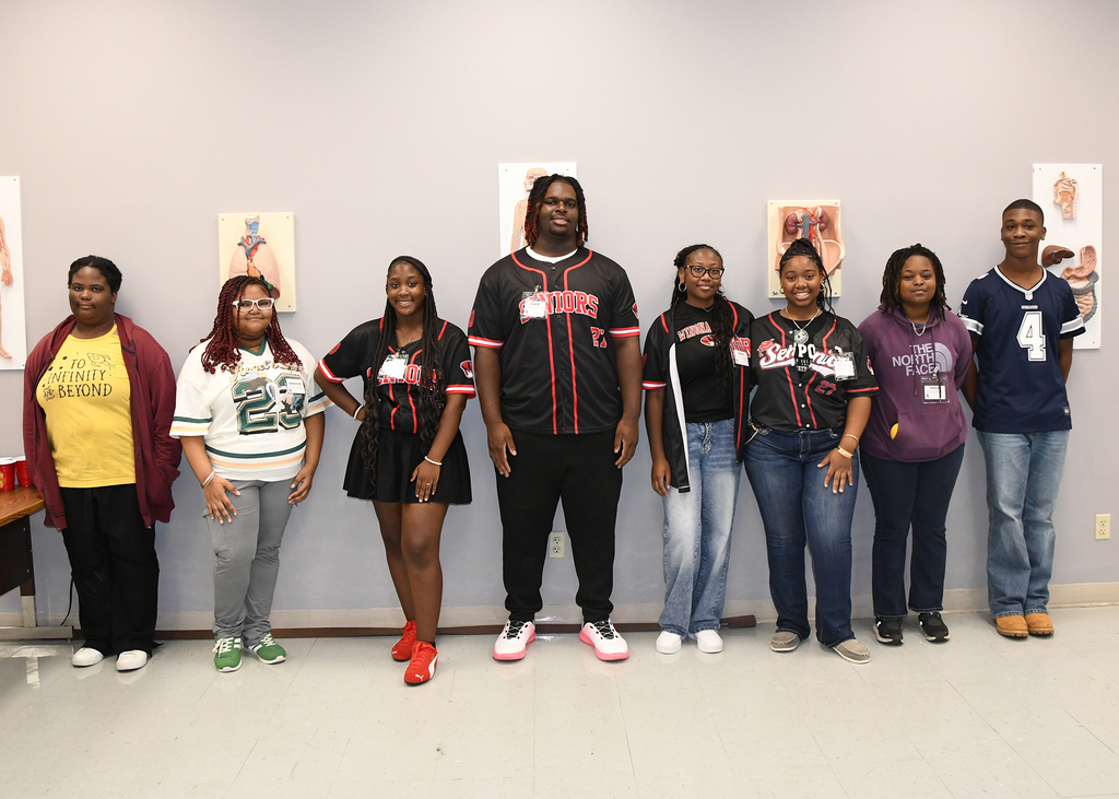 Nine individuals stand in a row against a white wall, possibly for a group photo. They wear various colors and styles of clothing.