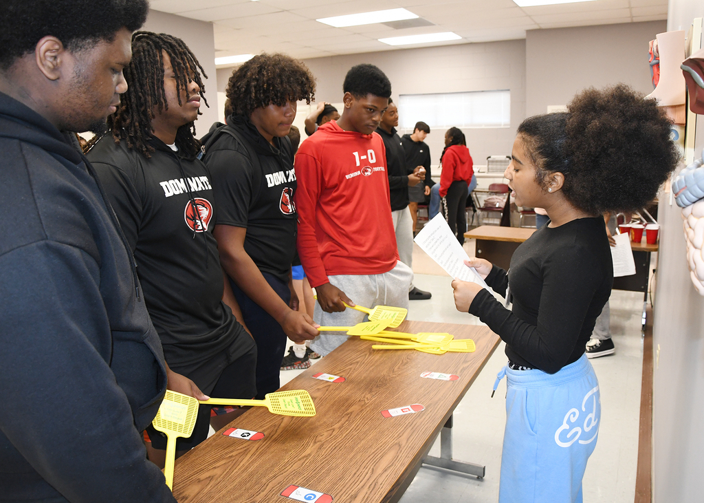 A group of people are gathered around a table with yellow items, a girl holds a paper.