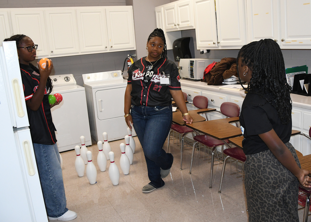 Three women in a room with a bowling set, refrigerator, and washer; one holds an orange.