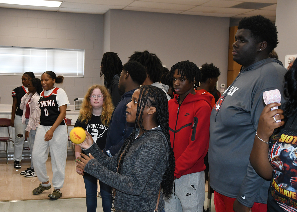 A group of people standing in a room; one holds a yellow ball and speaks to others.