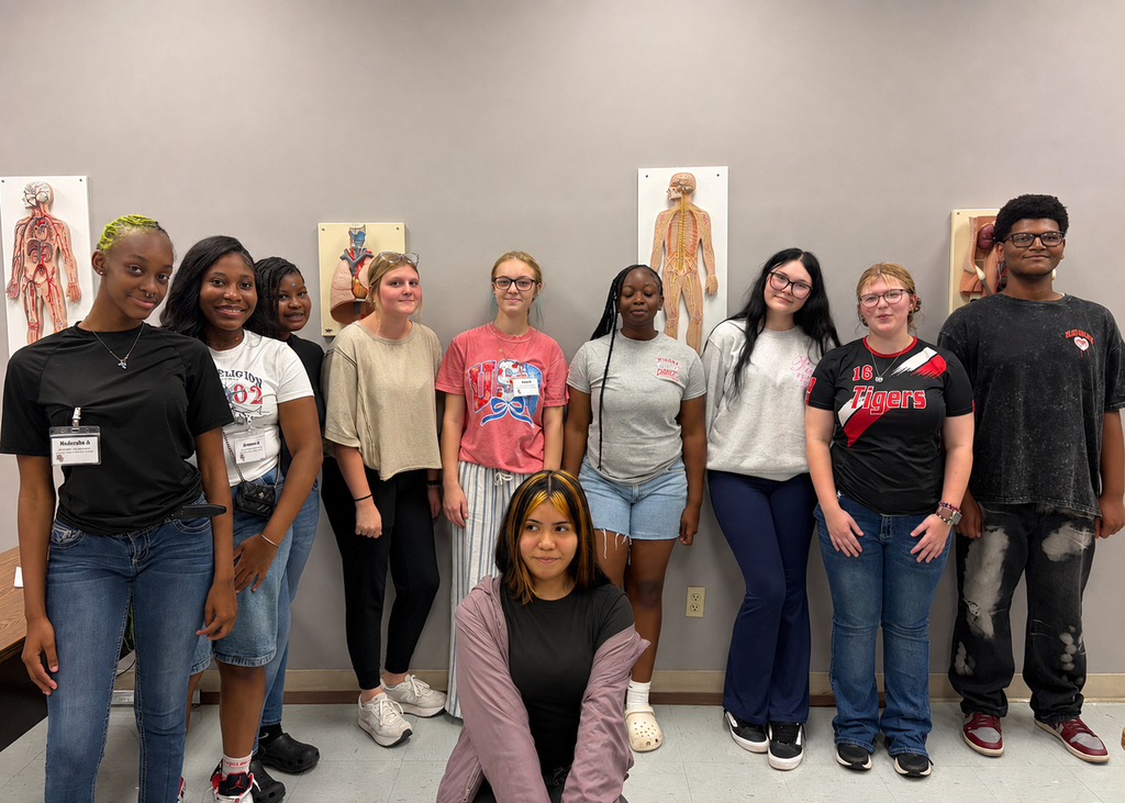 A group of students stands in a room with a white wall. Some wear sneakers, others sandals. Posters line the wall behind them.