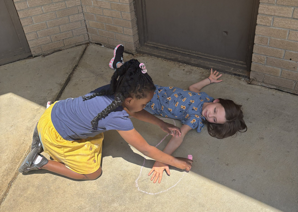 Two children, one kneeling and one lying on their back, play on a concrete floor, drawing with chalk.