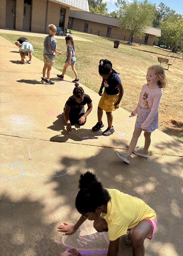 Children on a concrete path draw with chalk. One child kneels, others stand. Building and trees in background.