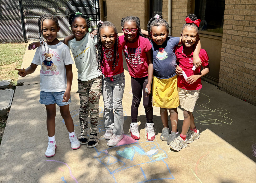 Five girls pose for a photo outside on a sidewalk with chalk drawings; one holds a white object.