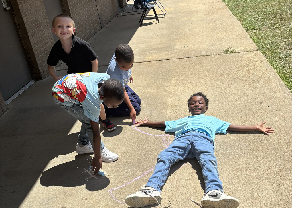 Four children playing outside; one lying on the ground, three standing, one holding a chalk piece.