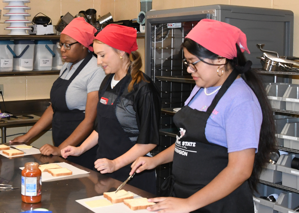 Four individuals in aprons and headscarves in a kitchen. One person slices bread on a cutting board.