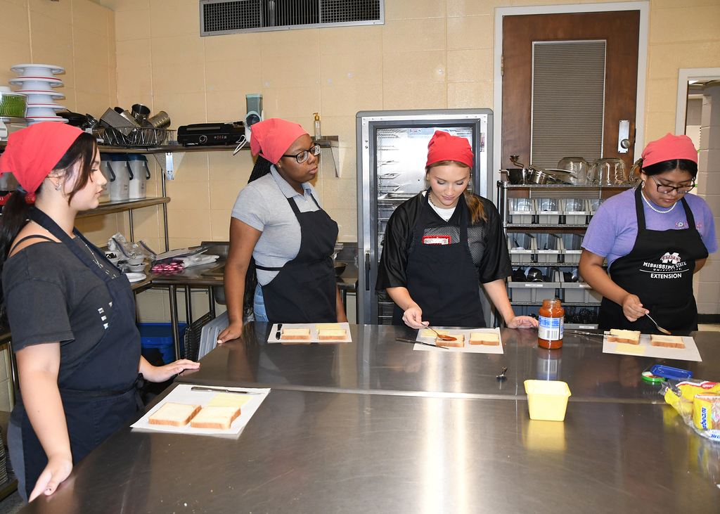 Five women with red headbands, wearing aprons, in a kitchen, preparing food on trays on a table.
