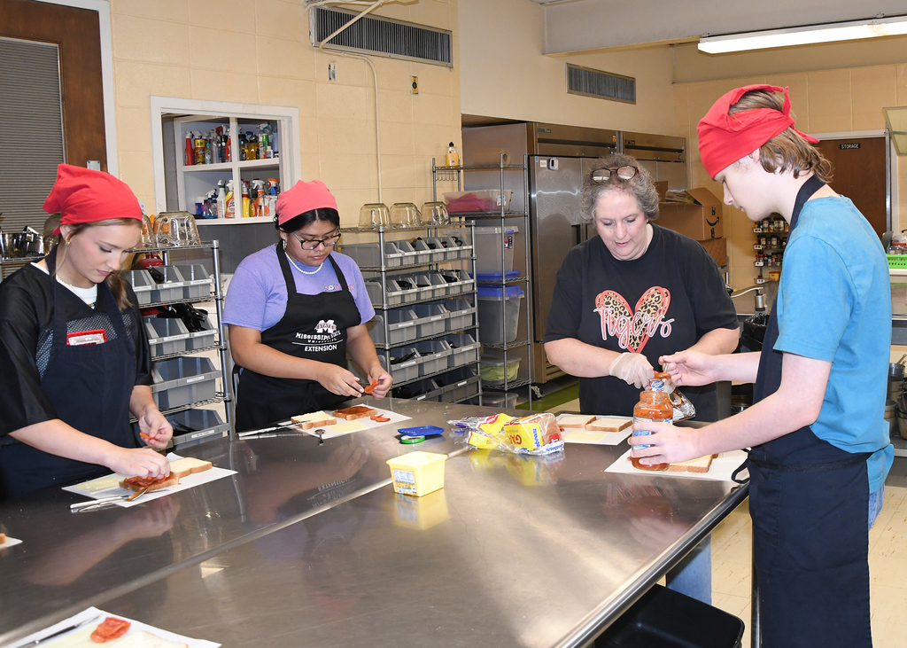 Several individuals with red hats wear aprons. They stand around a table and prepare food.