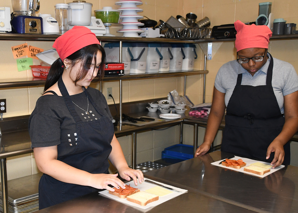 Two women in chef's hats and aprons prepare food in a commercial kitchen with stainless steel shelves and countertops.