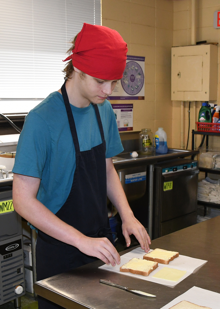 A person in a blue shirt, black apron, and red headband is preparing cheese sandwiches on a counter.