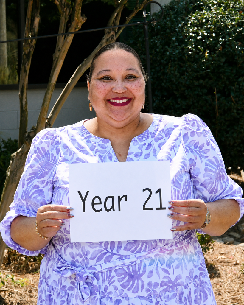 A woman in a purple dress holds a white sign reading "Year 21." She smiles and poses outside.