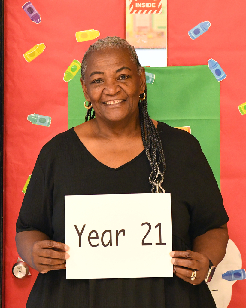 An adult woman smiling and holding a sign that reads "Year 21" in front of a colorful bulletin board.