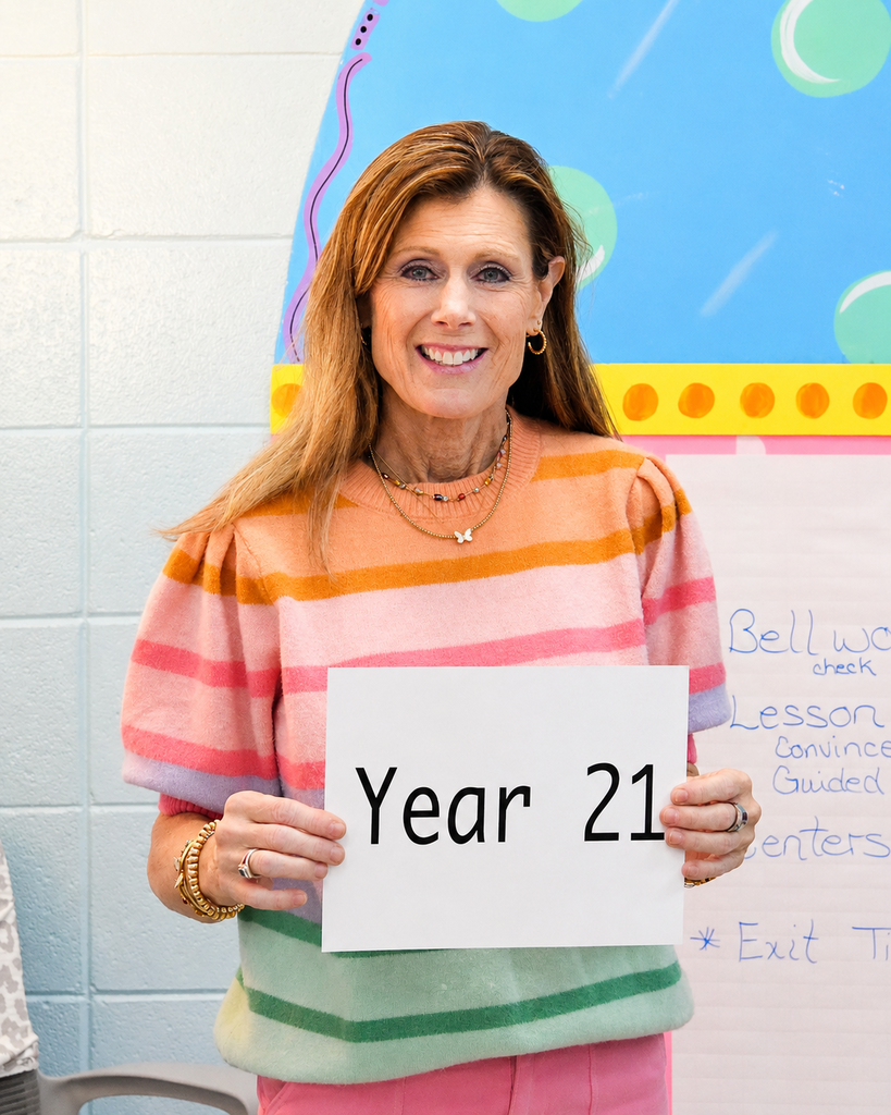 Woman in colorful striped shirt holds a sign reading "Year 21" in front of a colorful classroom background.