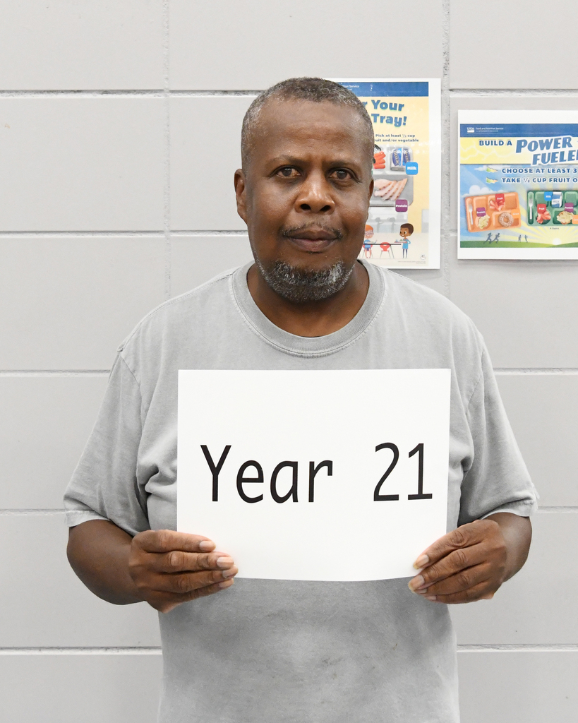 Man in gray shirt holding a white sign reading "Year 21" against a white background with posters.