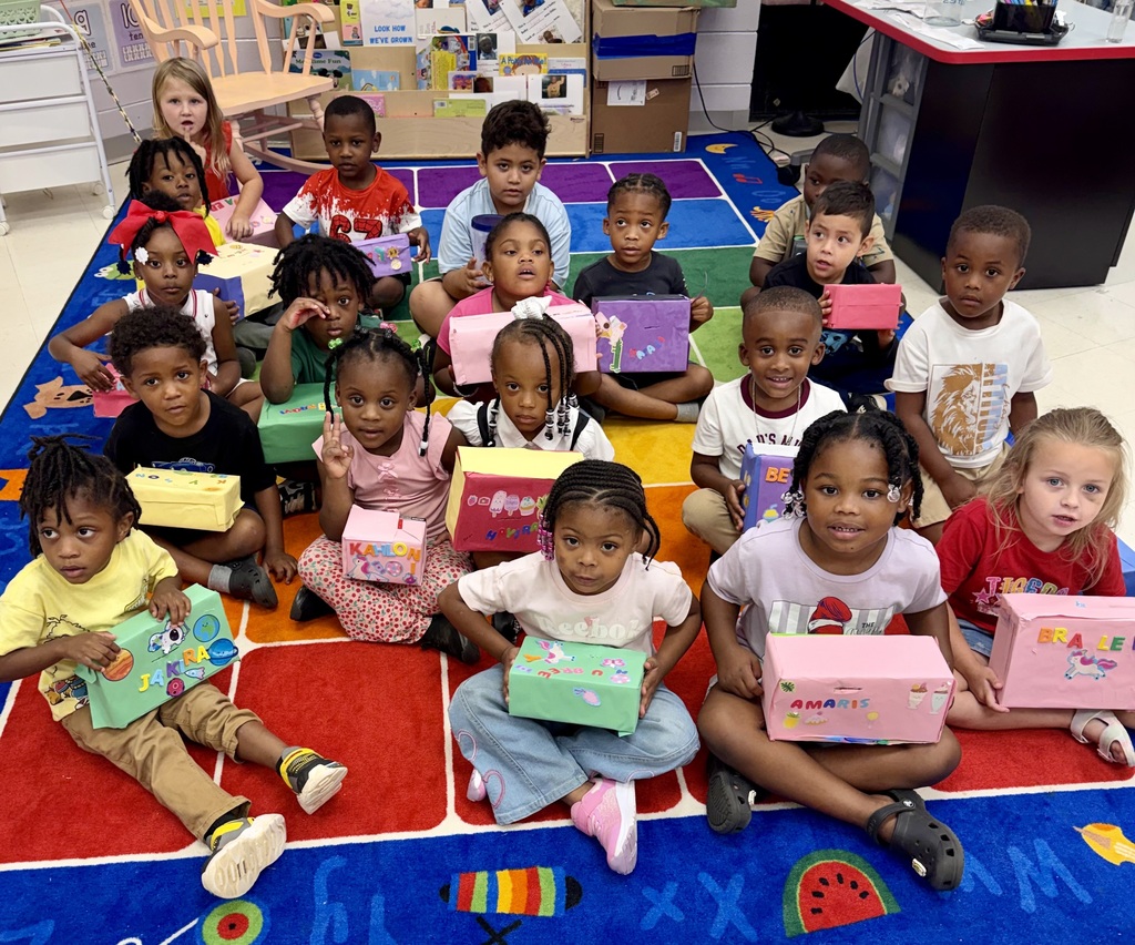 A group of children in a classroom holding gifts sit on a rug with a colorful design.