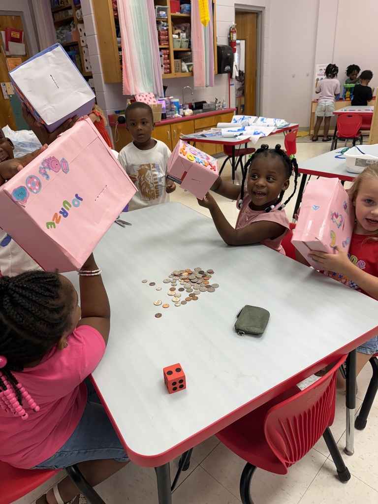 Five children sit around a table with coins and bags; one holds a dice. The room has white walls and red chairs.