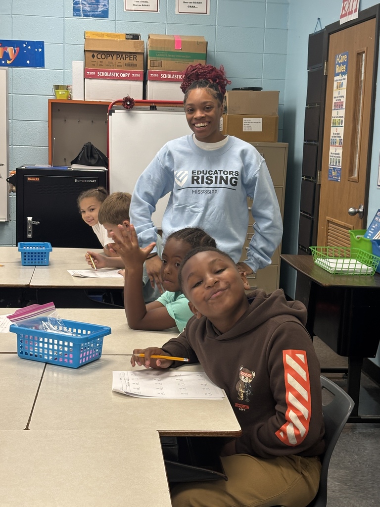 Two women and four children are in a classroom. One child is sitting at a desk, smiling.