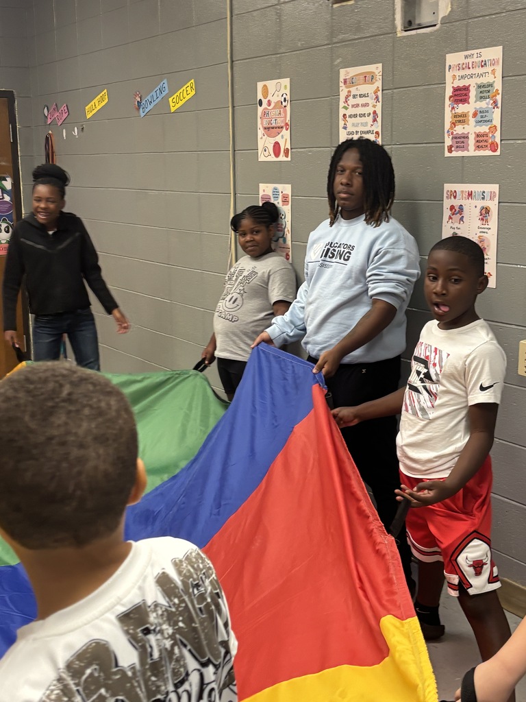 Four children in a classroom hold a colorful fabric while two adults stand behind them. Brightly colored posters adorn the wall.
