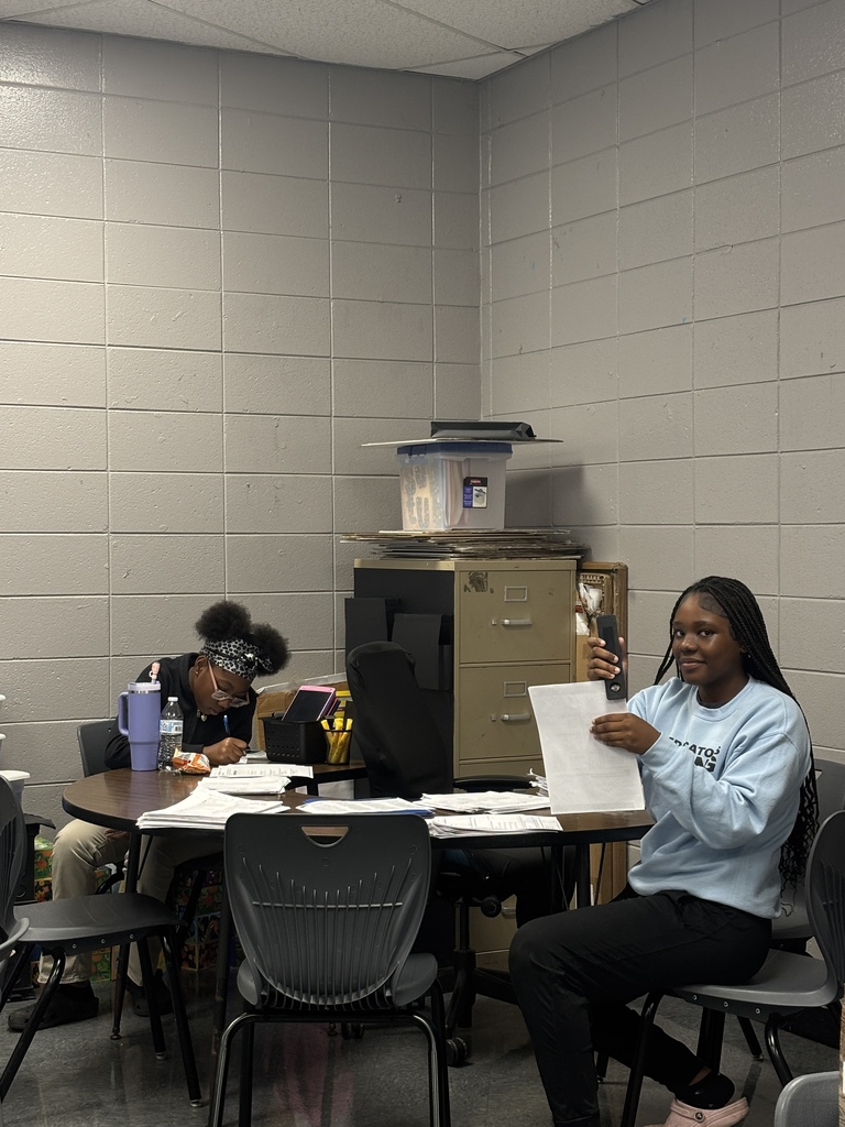 Two people in a room with chairs and a table. One holds papers and the other writes. Behind them is a storage cabinet and a shelf.