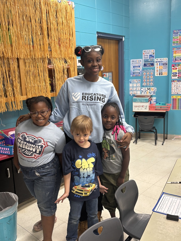 Four children stand together in a classroom. Two girls and two boys. A table and chairs are behind them.