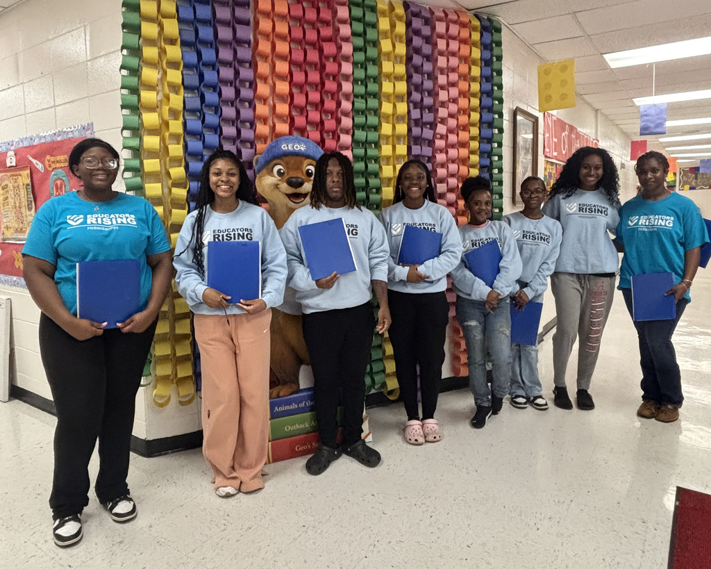 A group of students poses indoors; they hold blue folders and stand near a colorful paper chain wall.