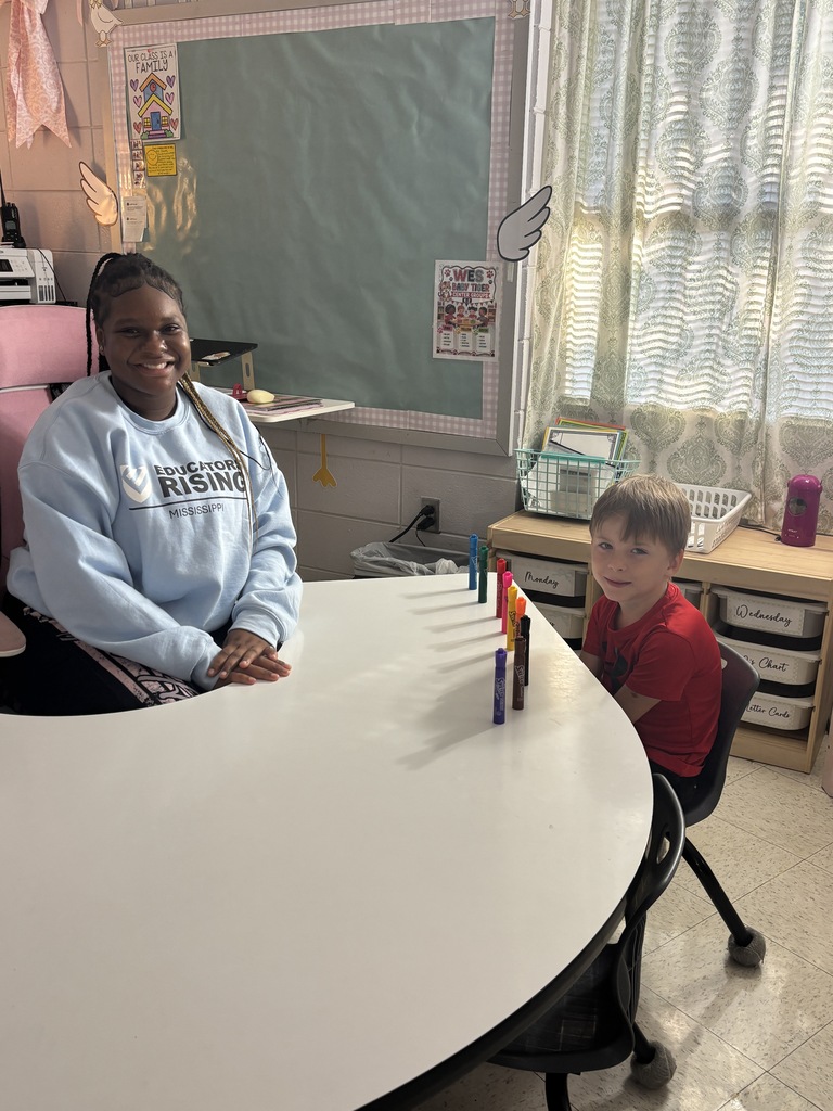 A woman and a boy sit at a round table with colored pencils, in a classroom with a white wall and window.