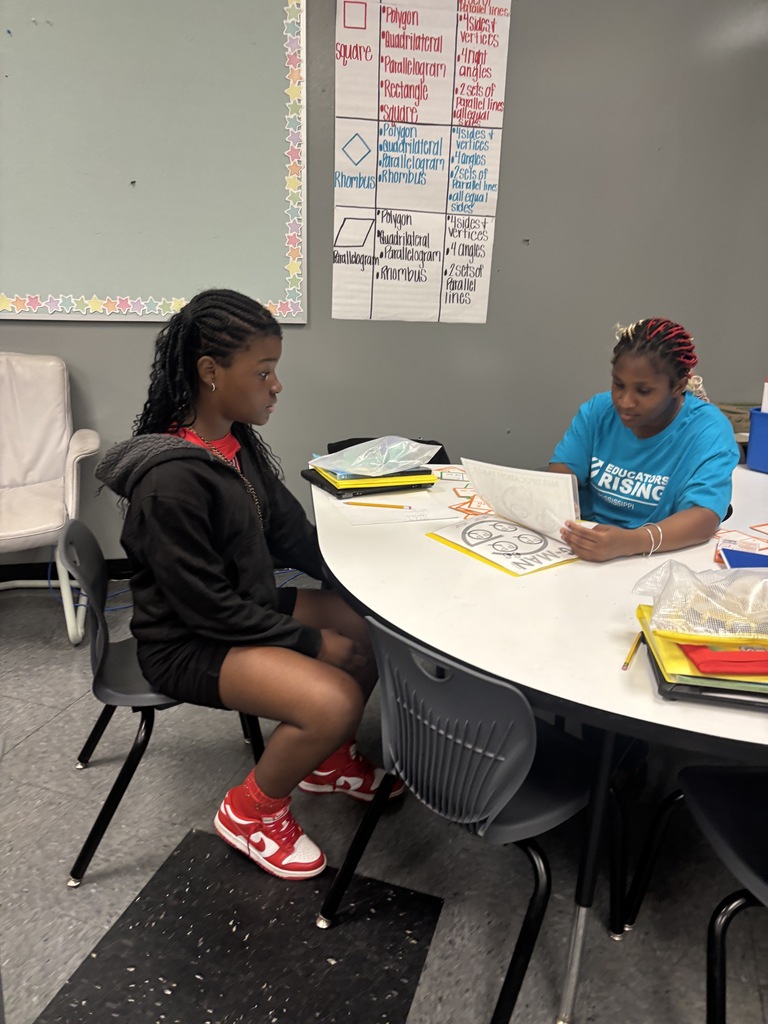Two students work at a table in a classroom, one with a laptop and the other with a book.