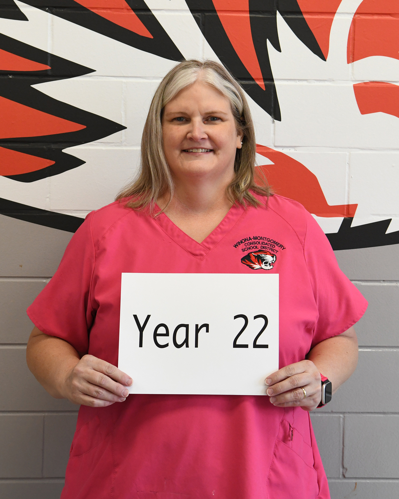Person in pink scrubs holding a sign that reads "Year 22" in front of a sports team logo.