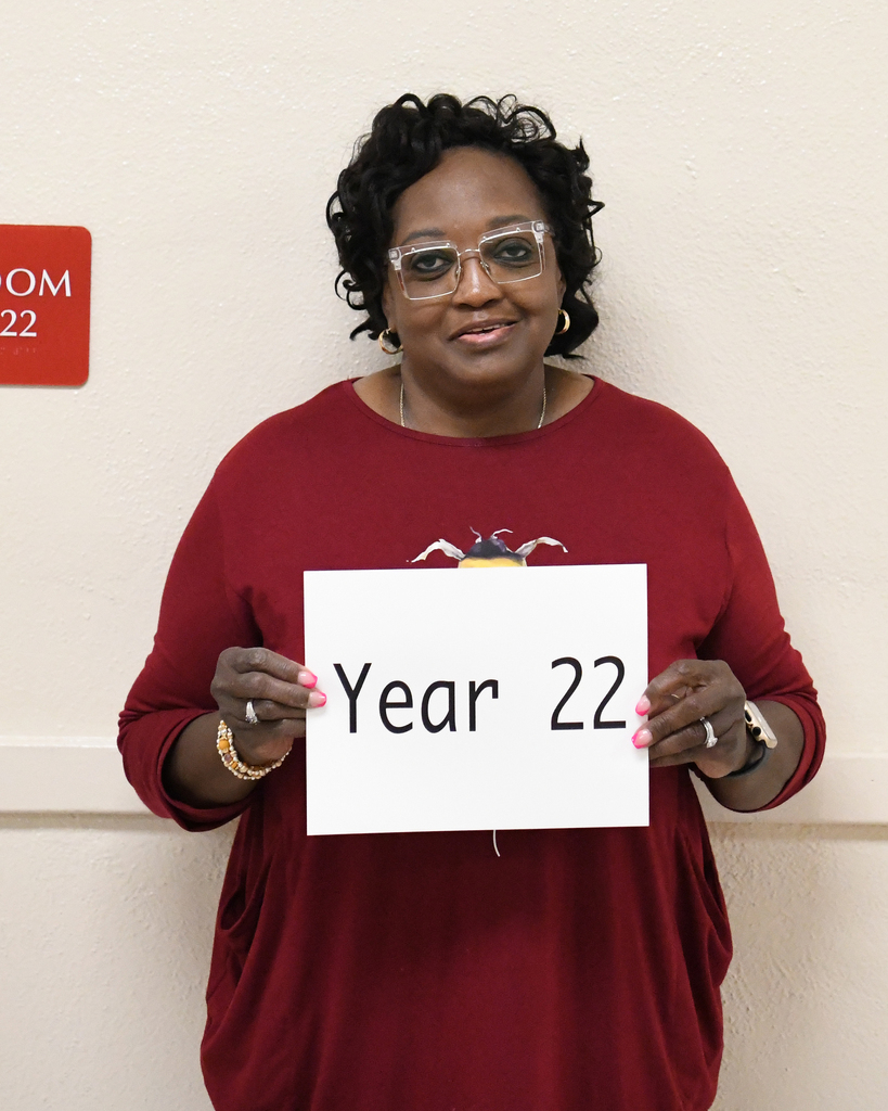Woman in red shirt holding a sign that says "Year 22" in front of a white wall.