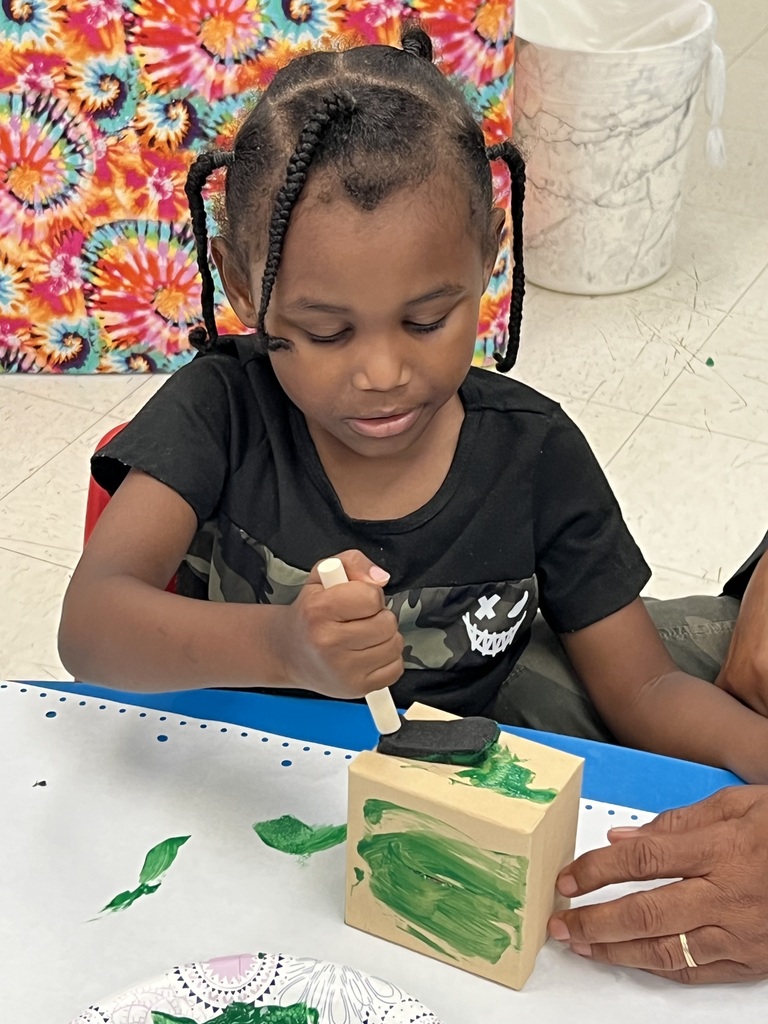 A girl with braided hair paints a wooden block with green paint at a table. A colorful backdrop is behind her.