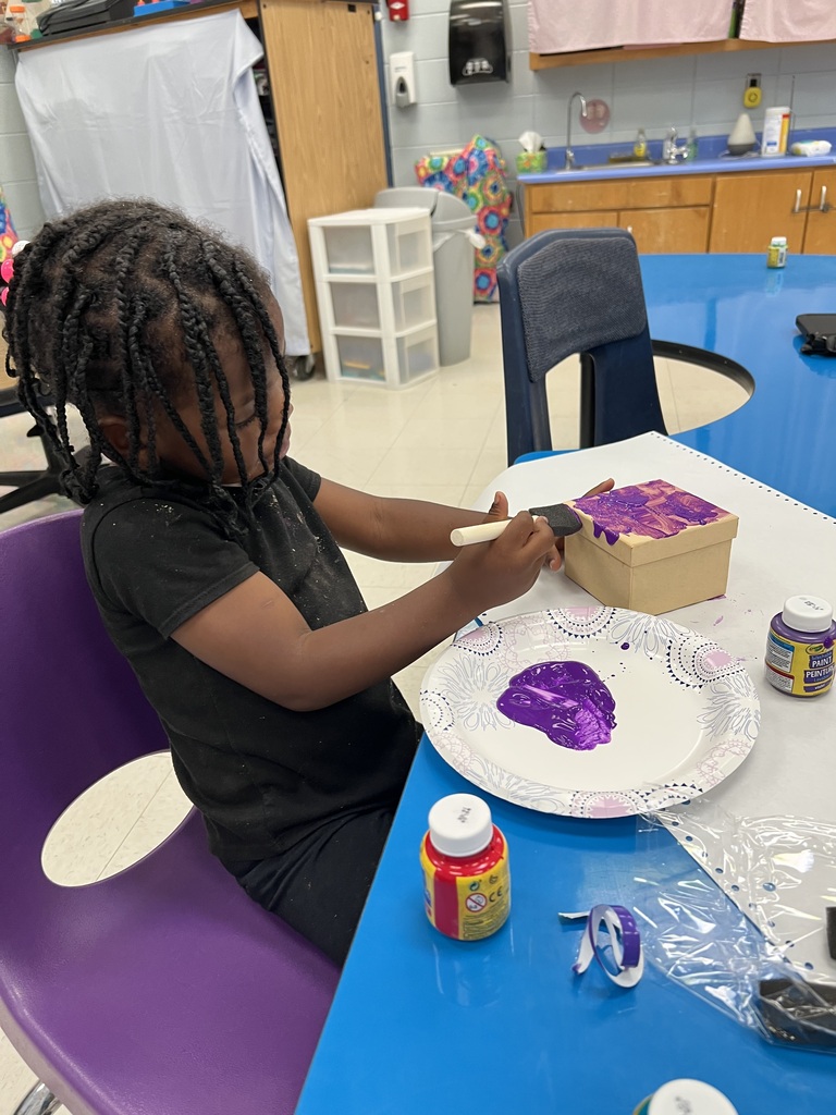 A child is seated at a table, painting purple paint on a box and paper.