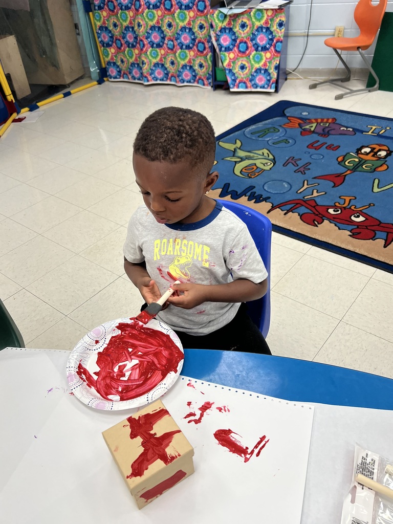 A child sits at a table, painting a red design on a paper plate and a box.