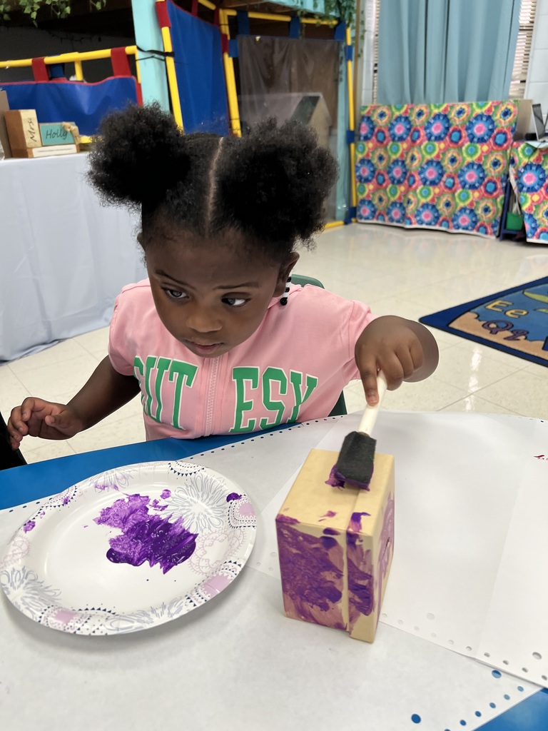 A young girl in a pink shirt sits at a table, painting a purple box. A paper plate with purple paint is beside her.