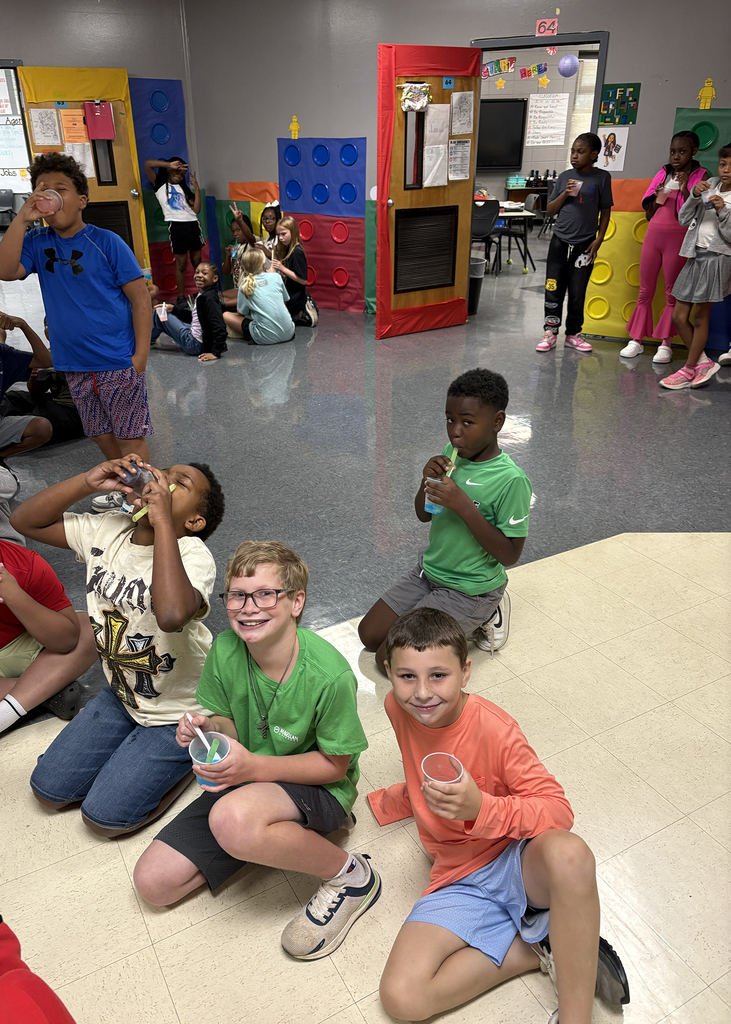 Children sit on the floor in a colorful classroom, holding drinks. One child sips from a plastic cup.