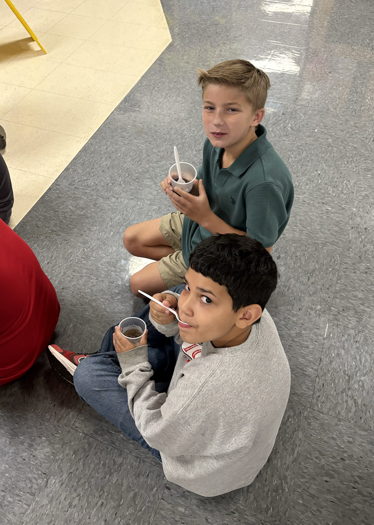 Two boys seated on the floor eating from plastic cups. One holds a spoon and the other holds a straw.