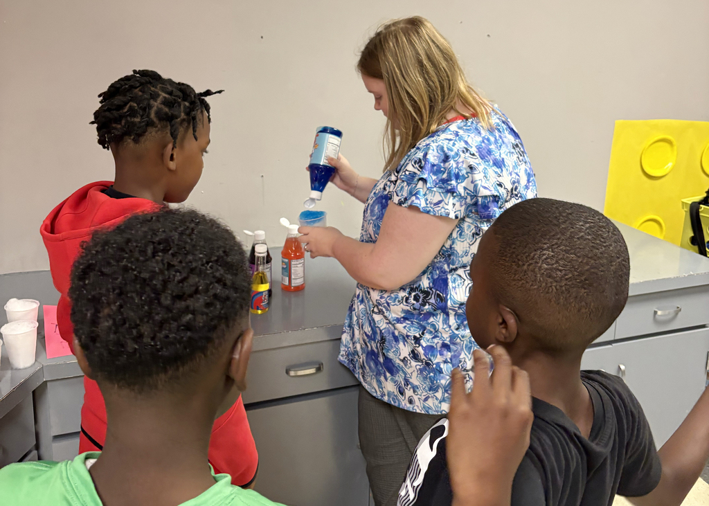 A woman in a blue floral shirt pours a liquid from a bottle into a cup while three children watch.