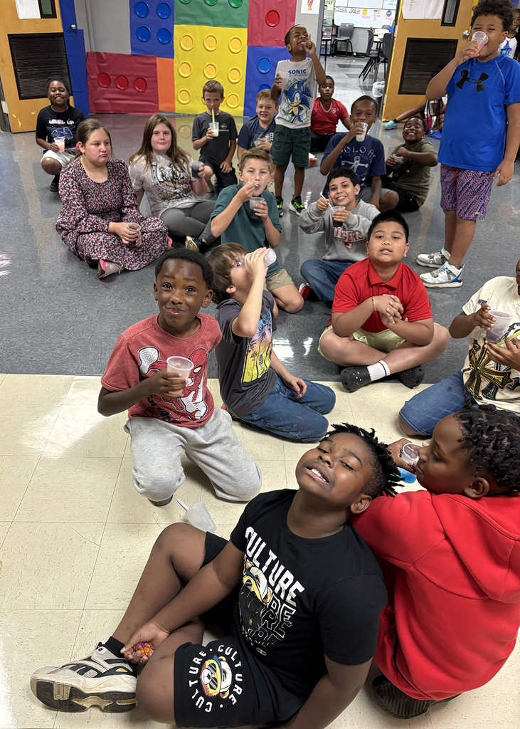Several children seated on the floor, some with drinks. One boy smiles, and some have food.