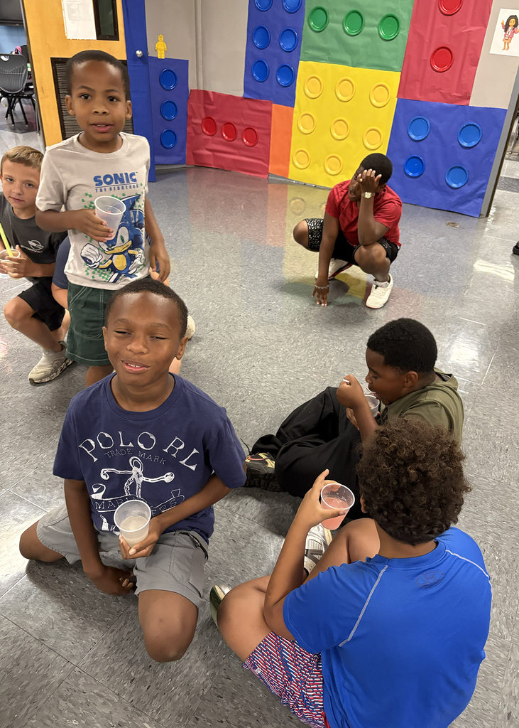Children sitting on the floor, one holding a cup, surrounded by colorful blocks. Bright colors in the background.