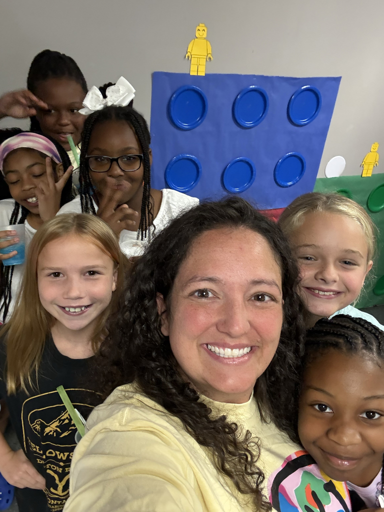 A woman with curly hair poses for a selfie with a group of children. They all smile. A blue structure in the background.