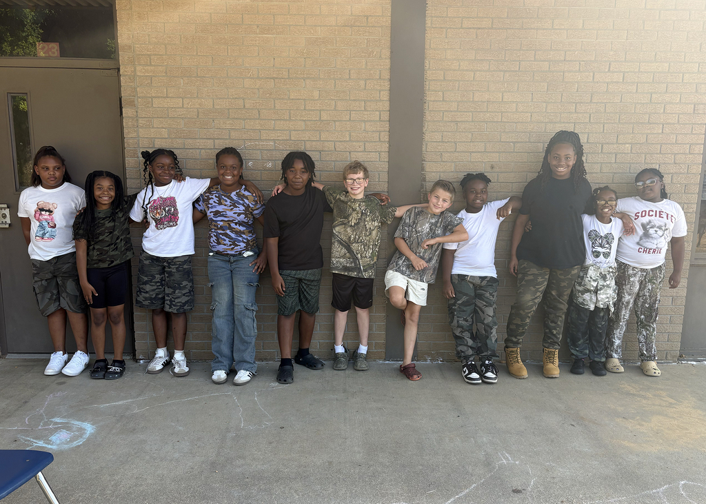 Group of children wearing various patterned shirts and pants. They stand in a line in front of a brick wall.
