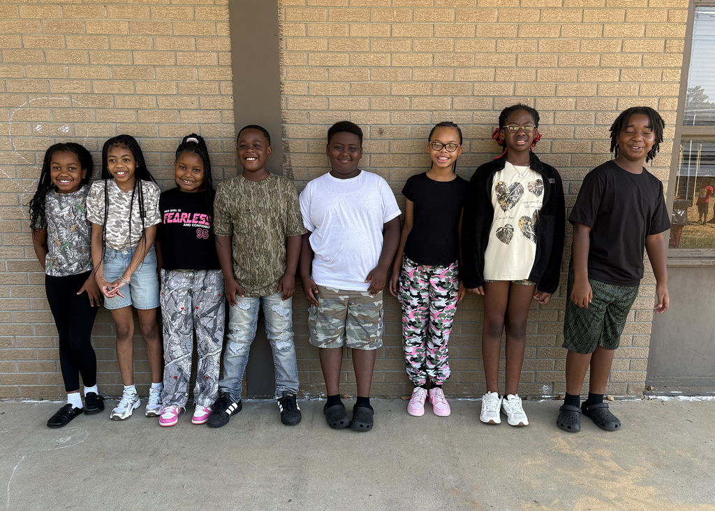A group of children stands in a line outside. Each child wears casual clothing and shoes. A brick wall is in the background.