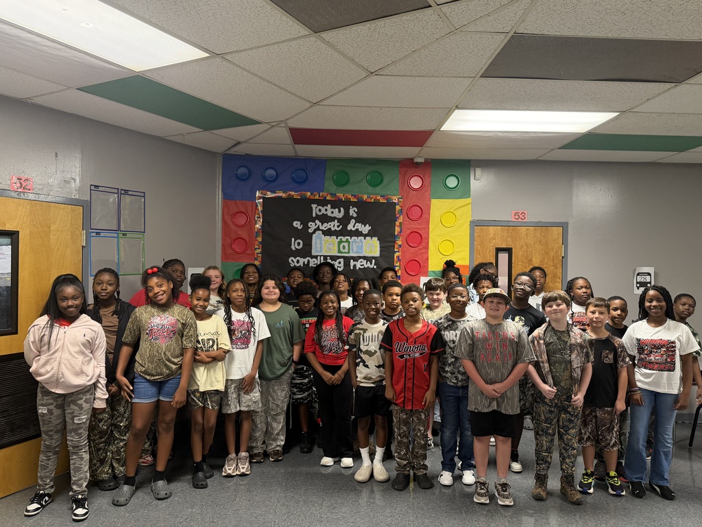 A group of children and a few adults stand in a room, with a bulletin board in the background.
